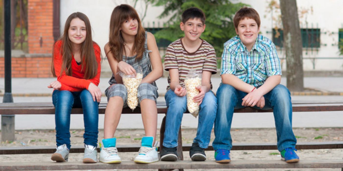 Zwei Mädchen und zwei Jungen, junges Teenageralter, sitzen auf der Lehne einer Holzbank, Füße auf der Bank; der Junge rechts ist übergewichtig