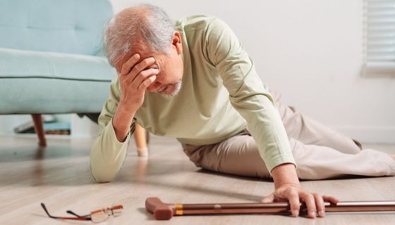 Ein älterer Herr ist gestürzt, er liegt auf dem Boden, bedeckt die Augen mit der Hand und greift nach dem Gehstock.