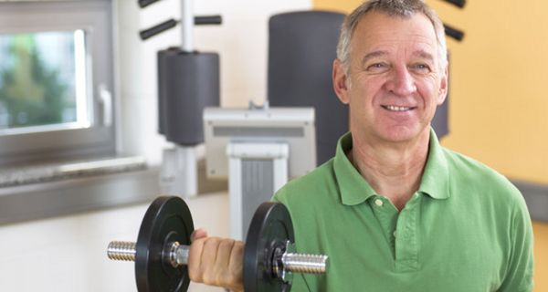 Mann, ca. Mitte 50, grünes Poloshirt, Frontalansicht, mit Hantel in der rechten Hand, gebeugter Arm, Hintergrund Fitnessgeräte im Studio