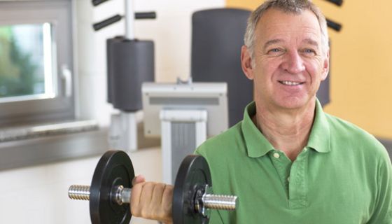 Mann, ca. Mitte 50, grünes Poloshirt, Frontalansicht, mit Hantel in der rechten Hand, gebeugter Arm, Hintergrund Fitnessgeräte im Studio