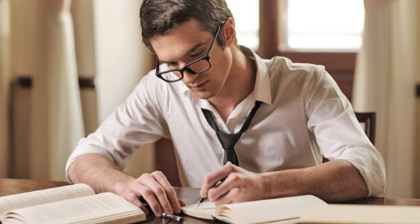 Junger Mann mit Brille, Hemd und Krawatte sitzt an einem Tisch und schreibt mit der linken Hand einen Text