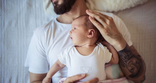 Vater, liegt mit seinem Baby auf dem Bauch auf einem Bett.