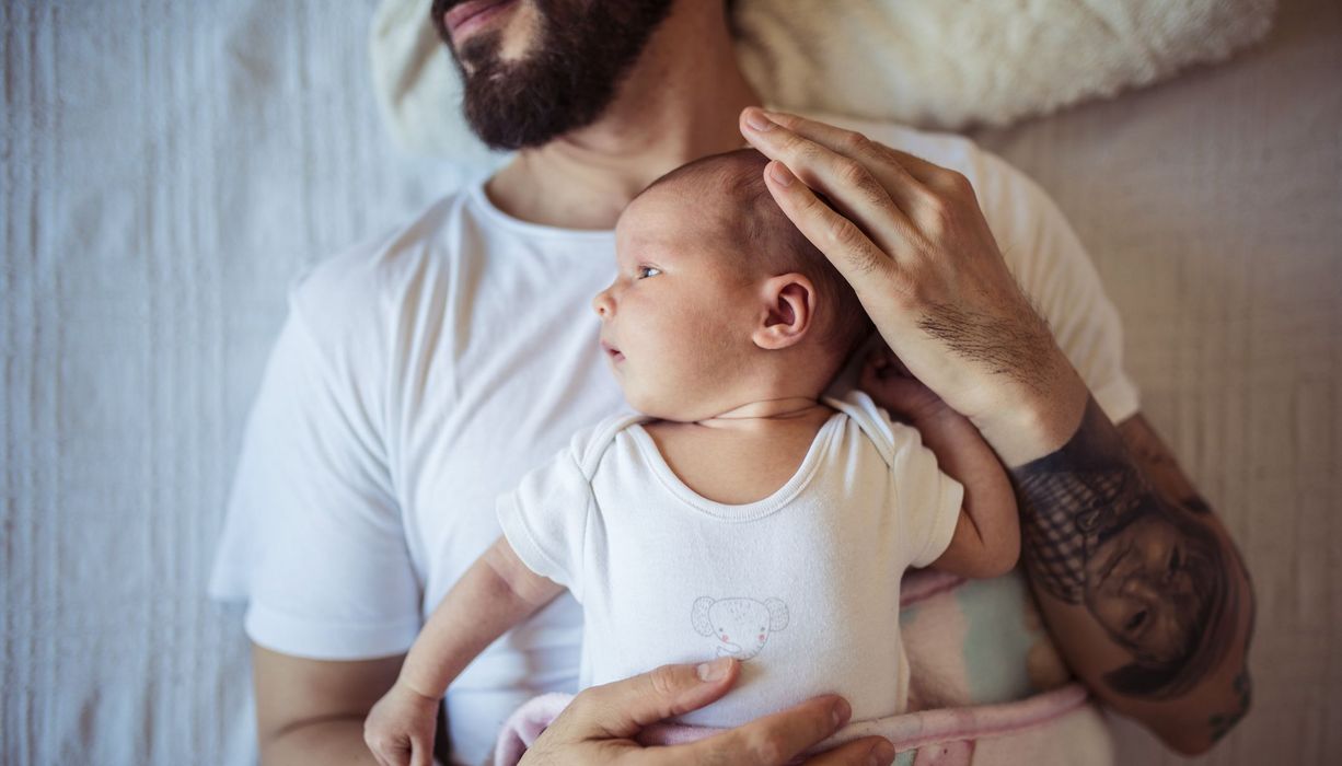 Vater, liegt mit seinem Baby auf dem Bauch auf einem Bett.