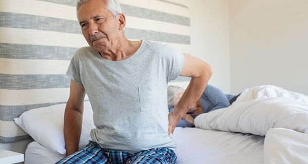Älterer Mann mit Kreuzschmerzen, sitzt auf seinem Bett.