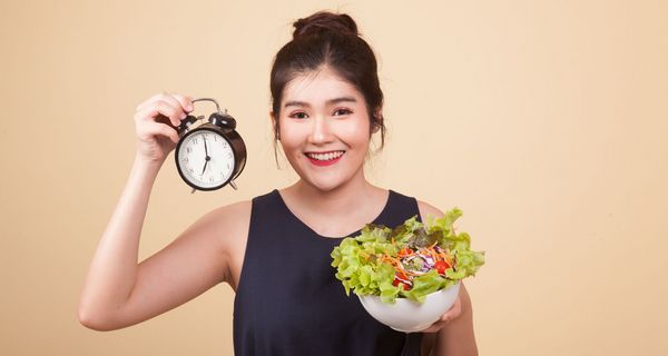 Eine Frau steht vor einem beigen Hintergrund. Sie hält eine Uhr in der rechten Hand und einen Salat in der linken Hand.