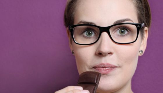 Portraitfoto junge Frau mit großer schwarzrandigen Brille schaut mit aufgerissenen Augen in die Kamera, ein Stück dunkle Schokolade zum Mund führend