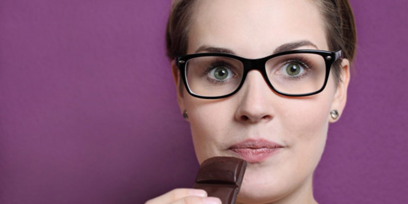 Portraitfoto junge Frau mit großer schwarzrandigen Brille schaut mit aufgerissenen Augen in die Kamera, ein Stück dunkle Schokolade zum Mund führend