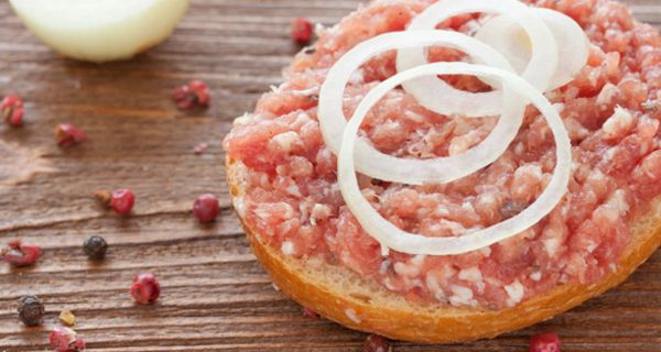 Halbes Mettbrötchen mit Zwiebelringen auf Holztisch, bunte Pfefferkörner, im Hintergrund Zwiebel