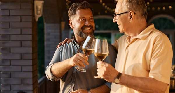 Ein älterer Herr und ein jüngerer Mann stoßen zusammen in der Abendsonne auf der Terrasse mit Wein an.
