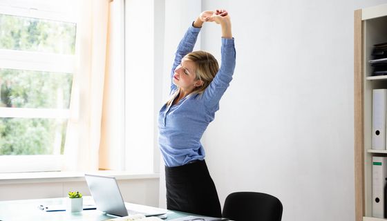 Frau in Bürokleidung steht vor einem Schreibtisch mit Laptop und streckt sich. 