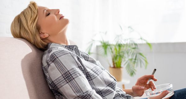 Eine Dame sitzt auf dem Sofa mit nach hinten gelehntem Kopf und hält einen Joint in der Hand.