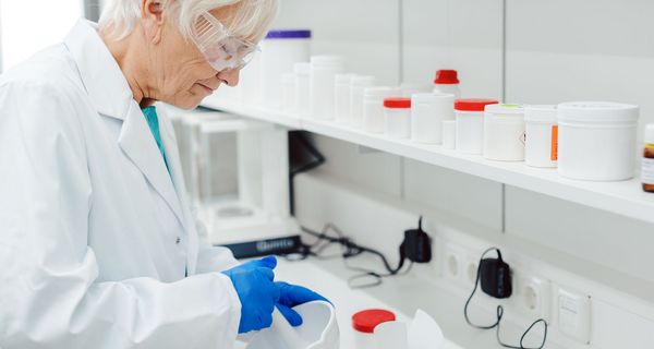 Eine Apothekerin mit Schutzbrille und -handschuhen mischt mittels eines Mörsers eine Apotheken-Rezeptur im Labor an.