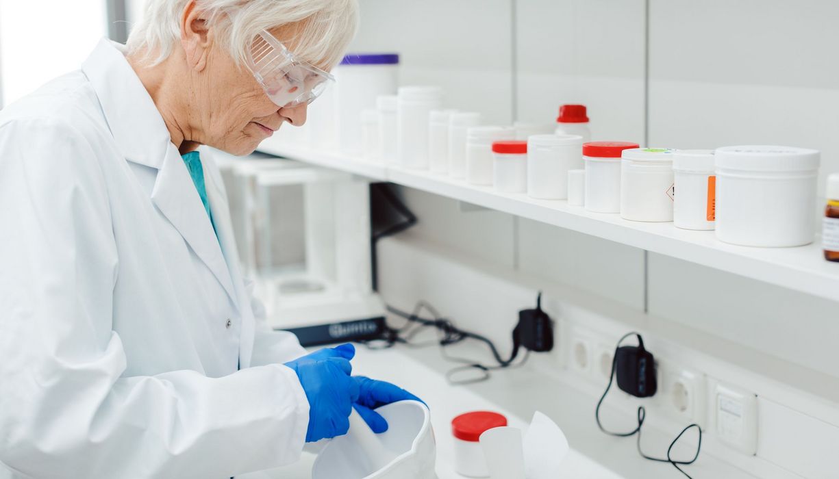 Eine Apothekerin mit Schutzbrille und -handschuhen mischt mittels eines Mörsers eine Apotheken-Rezeptur im Labor an.