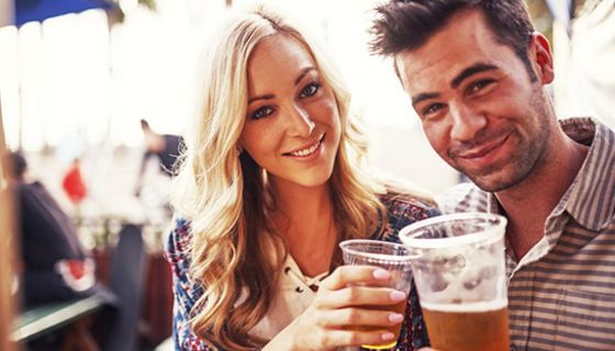 Paar, Ende/Mitte 30, draußen unter Sonnenschirm auf Fest (?), Frau blond, Mann dunkelhaarig, Freizeitkleidung, prosten sich mit Plastikbiergläsern zu, schauen dabei zur Kamera
