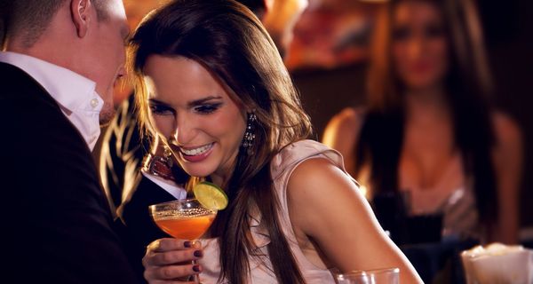 Ein Mann und eine Frau stehen bei einer Party gemeinsam an der Bar mti einem Cockteilglas in der Hand und flirten. 