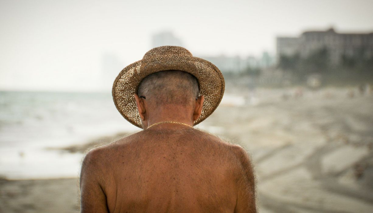 Senior mit freiem Oberkörper und Sonnenhut am Strand.