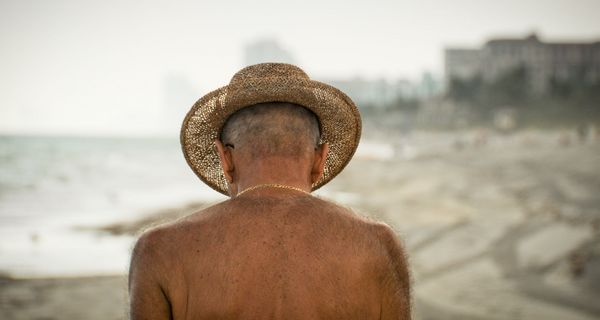 Senior mit freiem Oberkörper und Sonnenhut am Strand.