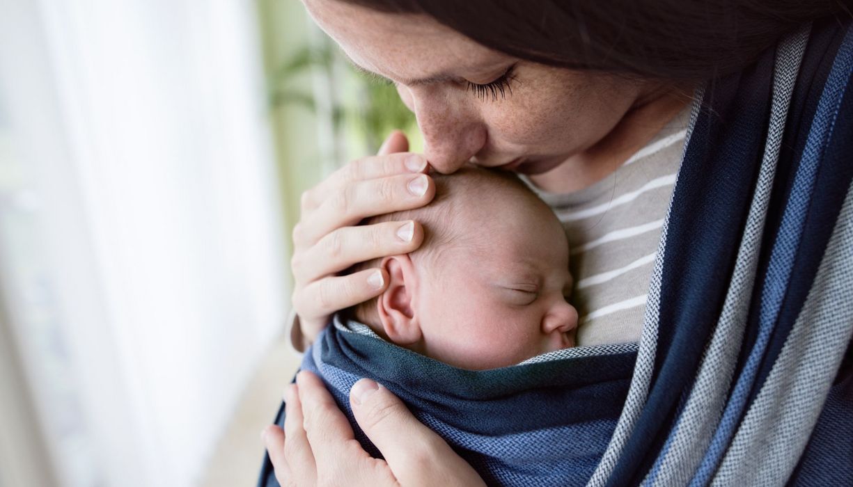 Mutter, hat ihr Baby in einer Trage und küsst es auf den Kopf.