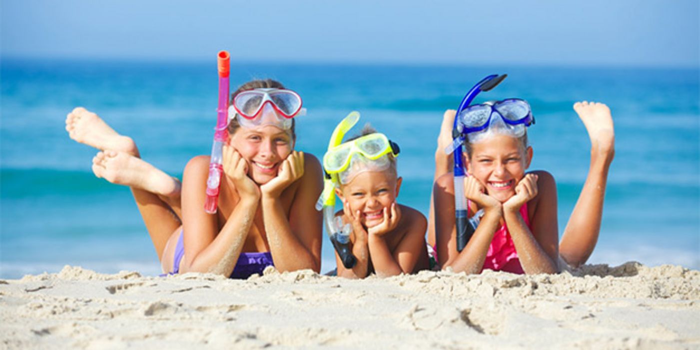 3 Kinder bäuchlinks am Strand, Badeklamotten, Taucherbrille, Schnorchel, Hände aufgestützt, in die Kamera lachend