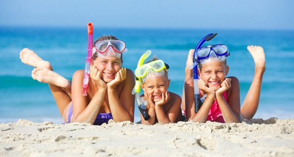 3 Kinder bäuchlinks am Strand, Badeklamotten, Taucherbrille, Schnorchel, Hände aufgestützt, in die Kamera lachend