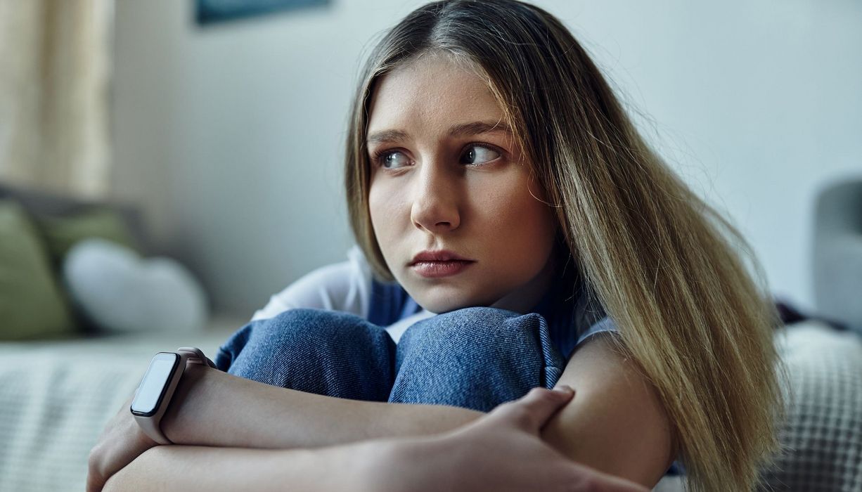 Eine Jugendliche sitzt am Fuß ihres Bettes, die Arme um ihre Kniee geschlungen, die Knie unter das Kinn gezogen. Sie blickt unsicher weg.