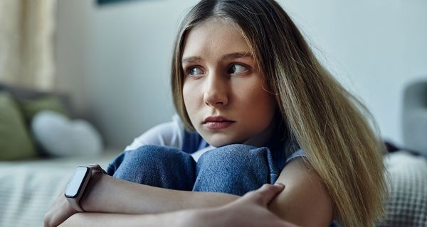 Eine Jugendliche sitzt am Fuß ihres Bettes, die Arme um ihre Kniee geschlungen, die Knie unter das Kinn gezogen. Sie blickt unsicher weg.