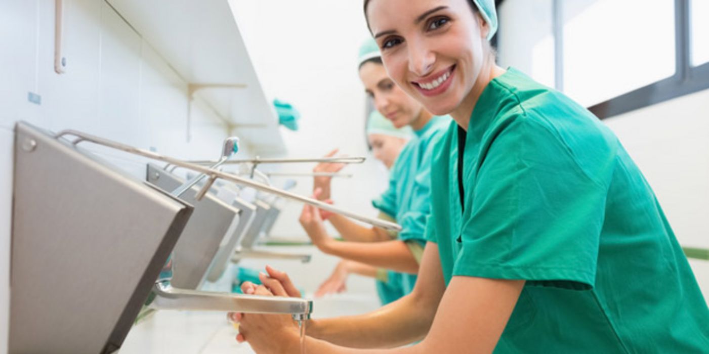 Krankenhauspersonal in OP-Kleidung beim Desinfizieren der Hände, im Vordergrund eine in die Kamera lächelnde junge Frau