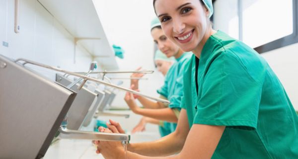 Krankenhauspersonal in OP-Kleidung beim Desinfizieren der Hände, im Vordergrund eine in die Kamera lächelnde junge Frau