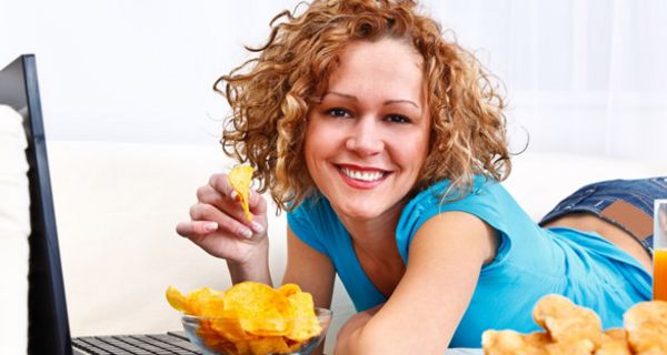 Junge Frau auf dem Bauch liegend vor dem Laptop mit einer Schüssel Kartoffelchips