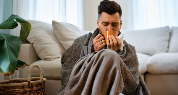 Mann, sitzt mit einer Decke und einer Tasse Tee auf der Couch.