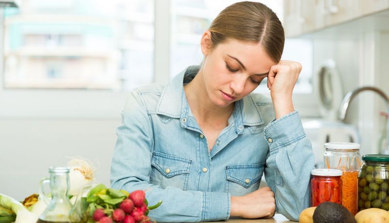 Müde, junge Frau stützt mit geschlossenen Augen ihren Kopf auf ihrer Hand ab. Auf dem Tisch vor ihr liegen Radieschen, Avocado, Karotte und Fenchel.