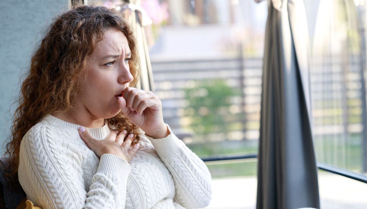 Eine Frau hustet und hält sich die Hand an das Schlüsselbein.