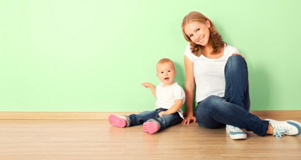 Teeniemutter, blond, Jeans, weißes Shirt sitzt  mit gleich gekleidetem blondem Kleinkind an pistazienfarbener Wohnungswand lächelt in die Kamera