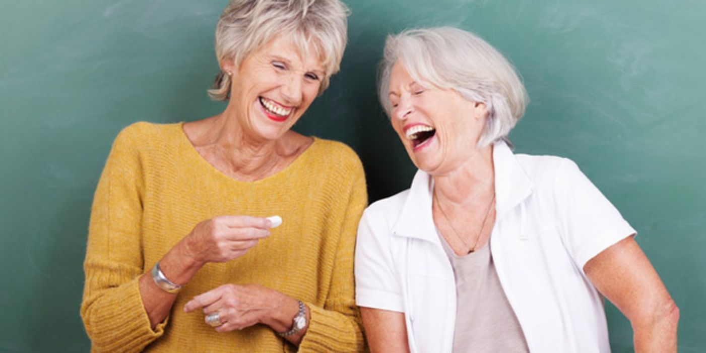 Zwei Frauen um die 60, 65 stehen lachend mit dem Rücken an einer Schultafel, die eine hält ein Stück Kreide in der Hand
