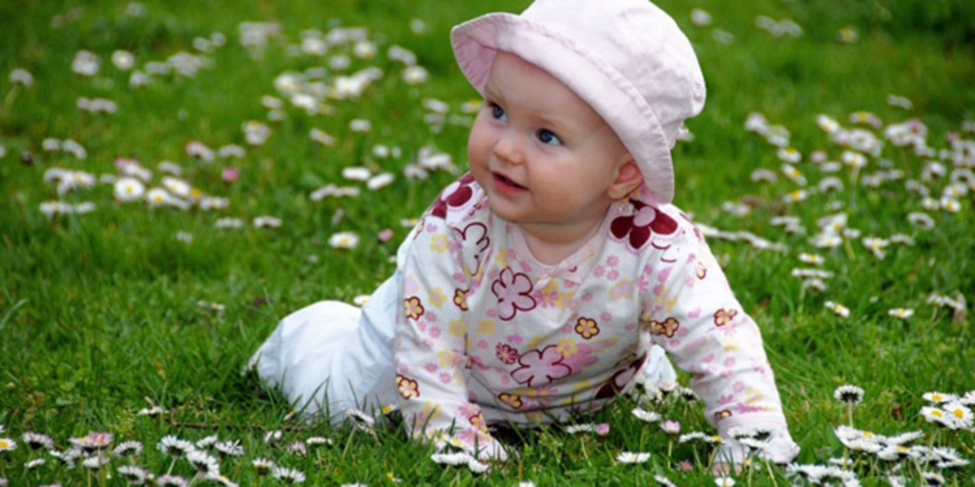 Freundlich blickendes Baby, ca. 12 Monate alt, auf Gänseblümchenwiese krabbelnd, weißes Hütchen, weißer Strampler mit rosa und zartlila Blütenmuster