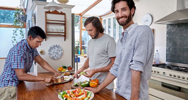 Drei Männer Ende 20 in Freizeitkleidung ín Küche beim Zubereiten einer vegetarischen Mahlzeit, bärtiger Mann im Vordergrund lacht in Kamera