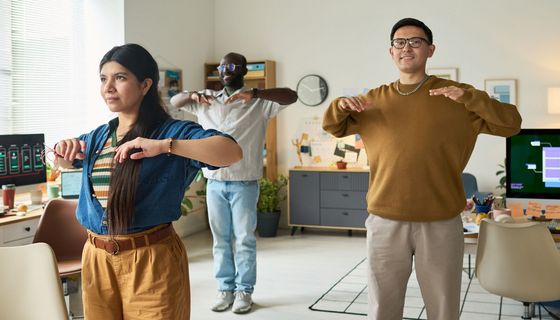 Eine Frau und zwei Männer machen einfache Fitnessübungen im Großraumbüro.