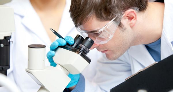 Junger Mann mit weißem Kittel und Schutzbrille schaut durch ein Mikroskop.