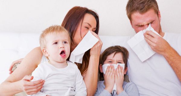 Mutter, Vater und ein Kind, ca 5 Jahre alt, sitzen im Bett und putzen sich die Nasen; ein ca. 3 jähriges Kind sitzt daneben und hustet