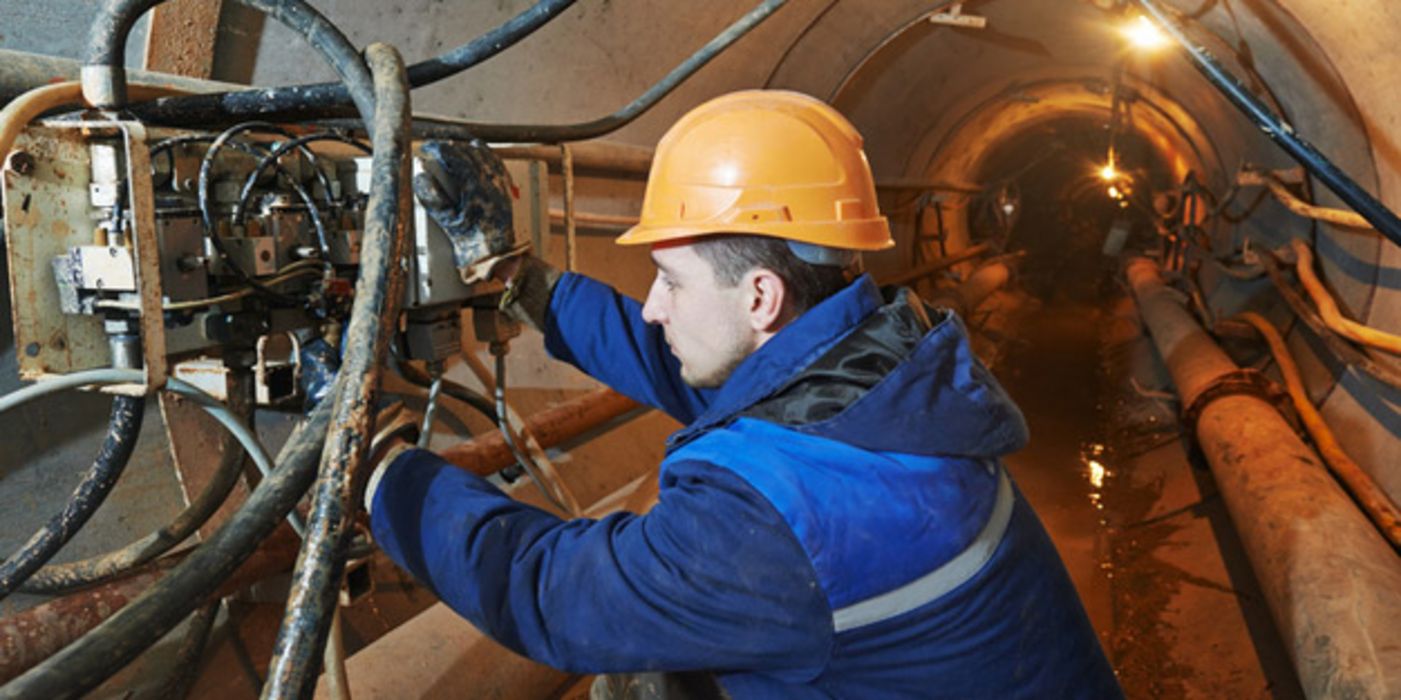 Mann um die 30 in blauer Arbeitskleidung, oranger Schutzhelm, unter Tage in einem Tunnel mit Rohrsystem bei der Arbeit