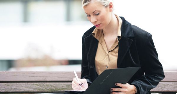 Frau in den 20ern, blonde, zurückgenommene Haare, dunkle Business-Jacke, helle Bluse, auf Bank sitzend in eine Bewerbungsmappe schreibend, ernster Gesichtsausdruck