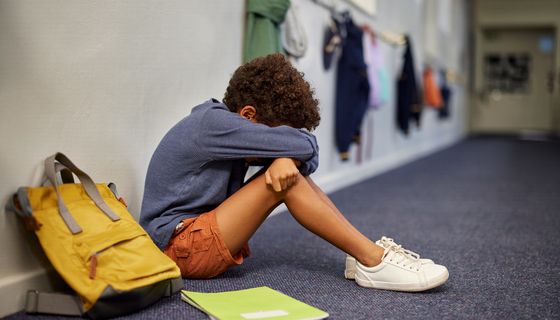 Trauriger Junge sitzt auf dem Flur in der Schule; neben ihm steht sein Rucksack, vor ihm liegt ein Heft auf dem Boden.