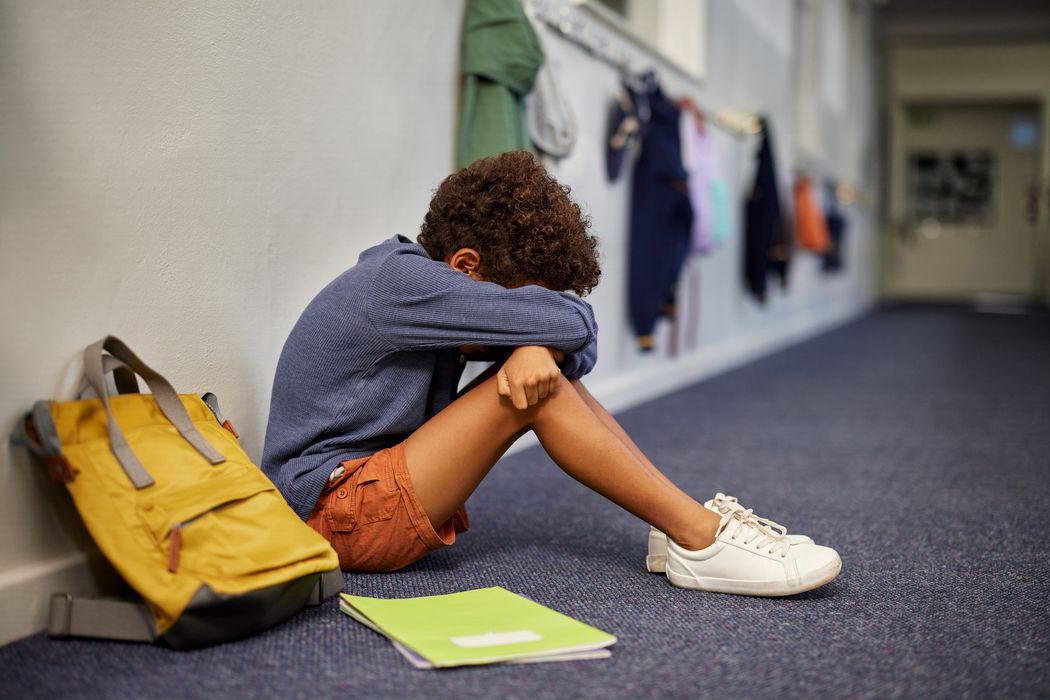Trauriger Junge sitzt auf dem Flur in der Schule; neben ihm steht sein Rucksack, vor ihm liegt ein Heft auf dem Boden.
