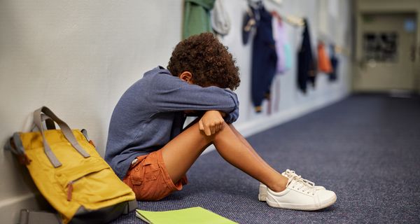 Trauriger Junge sitzt auf dem Flur in der Schule; neben ihm steht sein Rucksack, vor ihm liegt ein Heft auf dem Boden.