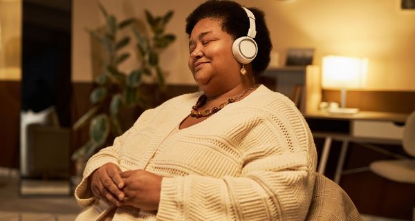 Eine Frau mittleren Alters sitzt auf einem Sessel. Sie lächelt und trägt Kopfhörer.