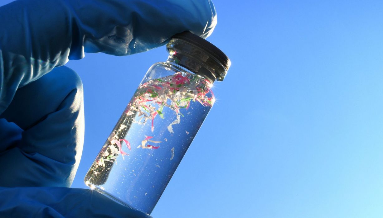Forscher mit Handschuh hält Ampulle mit Wasser zwischen den Fingern. Wasser ist sichtbar mit Mikroplastik verunreinigt.