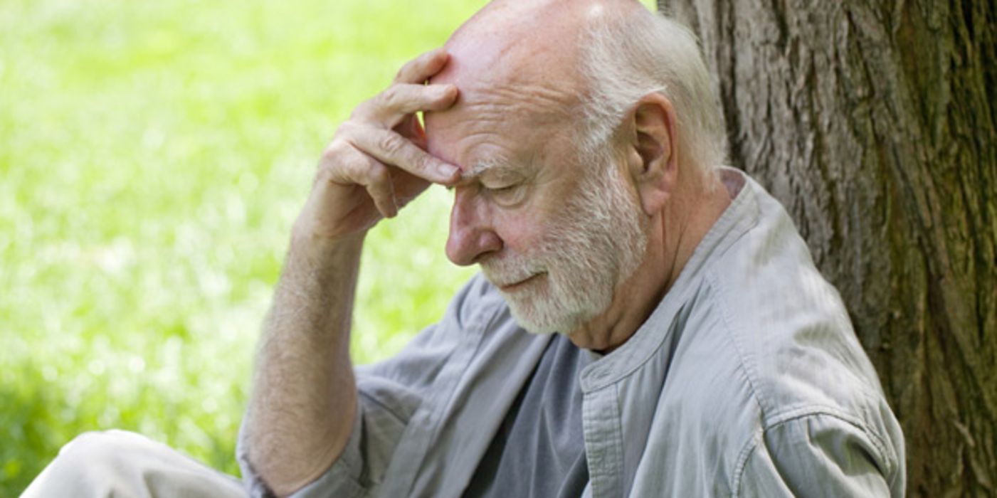 Grauhaariger Mann um die 70 mit Bart sitzt an einen Baum gelehnt und stützt den Kopf nachdenklich blickend in eine Hand