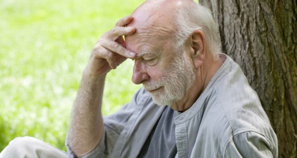 Grauhaariger Mann um die 70 mit Bart sitzt an einen Baum gelehnt und stützt den Kopf nachdenklich blickend in eine Hand