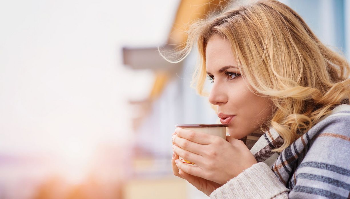 Blonde Frau mit Schal und Wollpullover, nippt an Kaffeetasse.