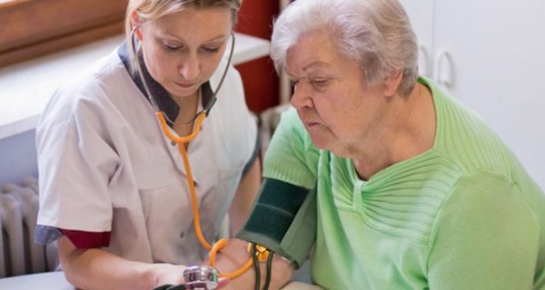 Alter Frau wird von junger Altenpflegerin der Blutdruck mithilfe von Oberarmmanschette und Stethoskop gemessen